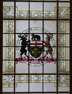 [White glass panels have a pattern outlined in gold and in the center is a crest with animals and a shield in it. These panels let natural light into the building.]